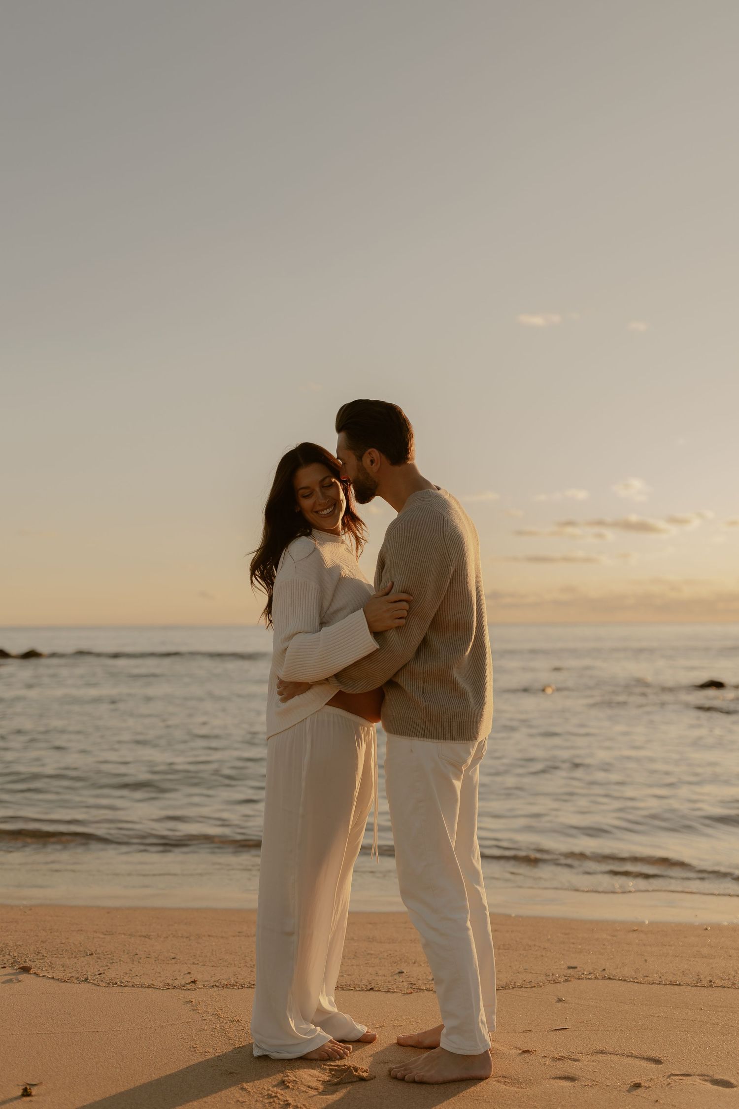 A couple in white outfits shares an intimate moment on a sandy beach at sunset.