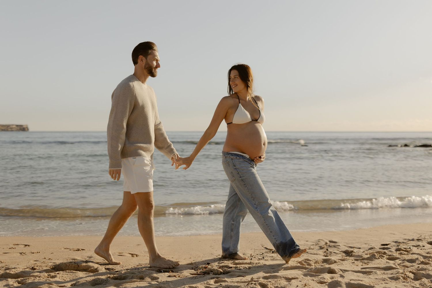 A couple walks hand in hand along the shoreline during sunset.