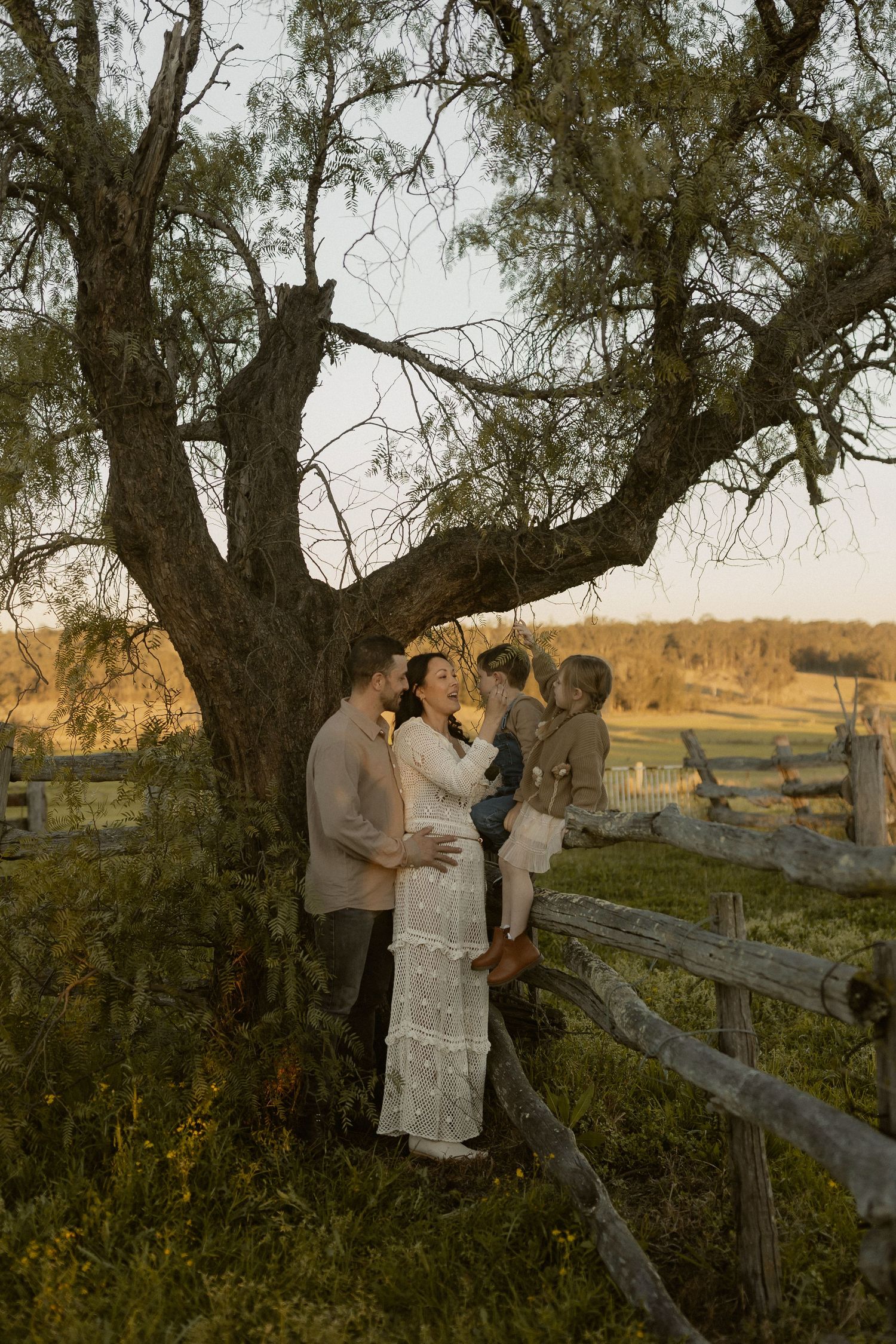Couple embraces beside a rustic wooden fence during golden hour in a countryside setting.