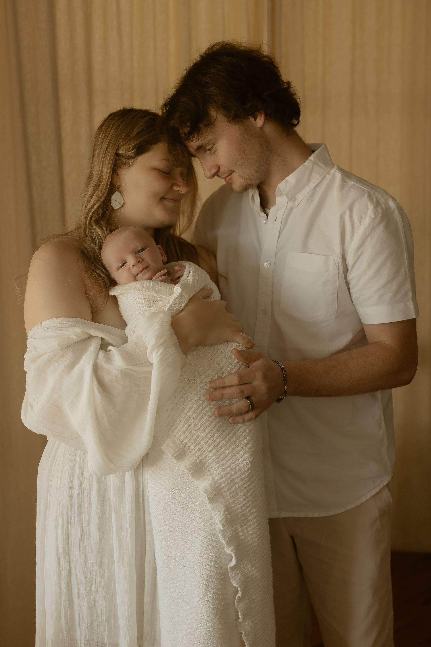 Family of three wearing white clothing embrace tenderly in a soft-lit indoor setting.