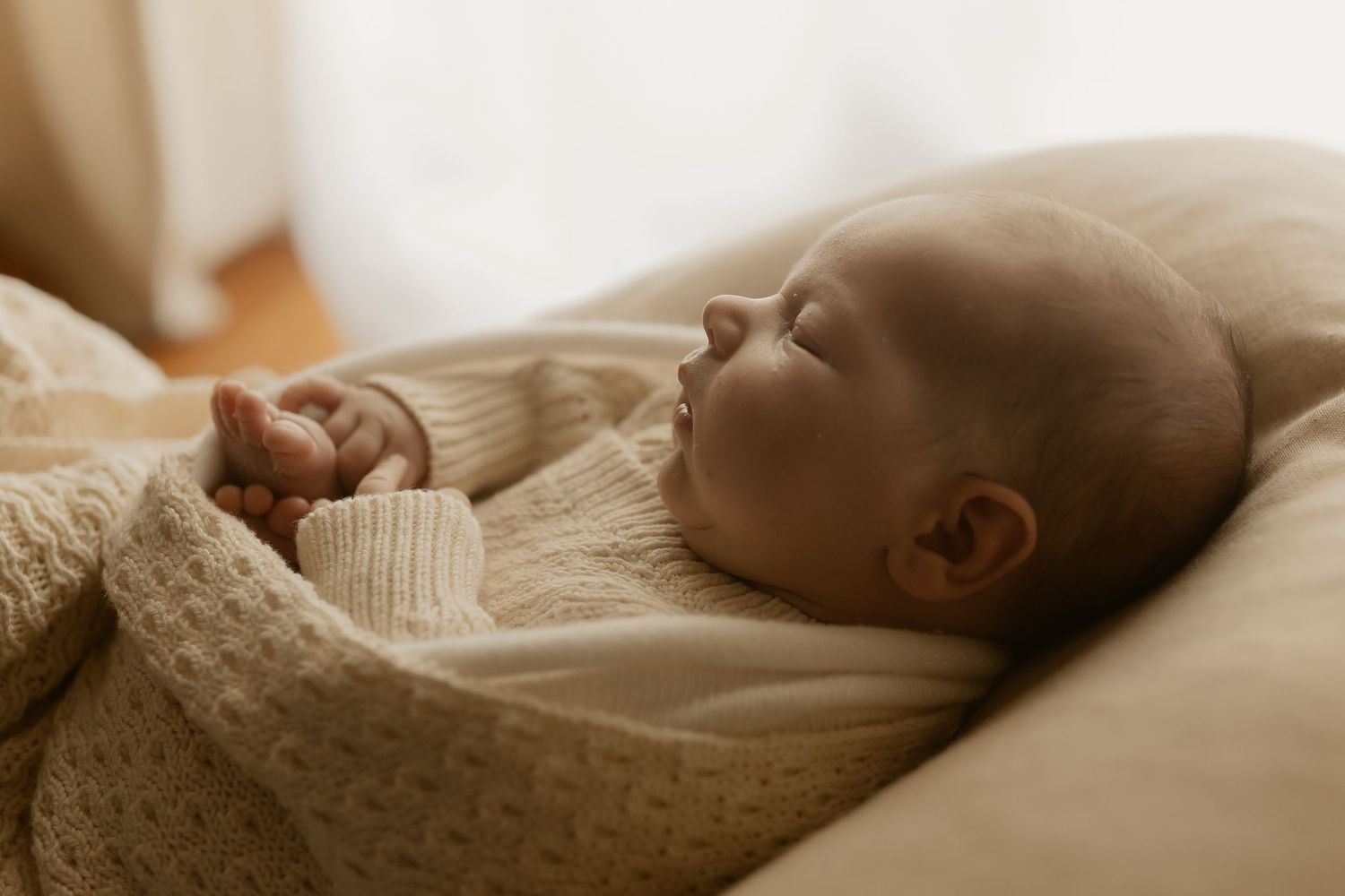 A sleeping newborn baby wrapped in a cream colored blanket rests peacefully against a light background.