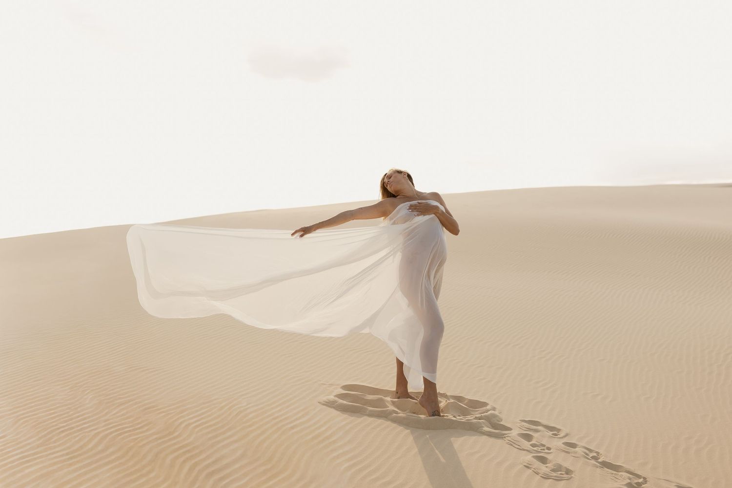 Person in flowing white dress poses dramatically across rippling sand dunes in warm sunlight.