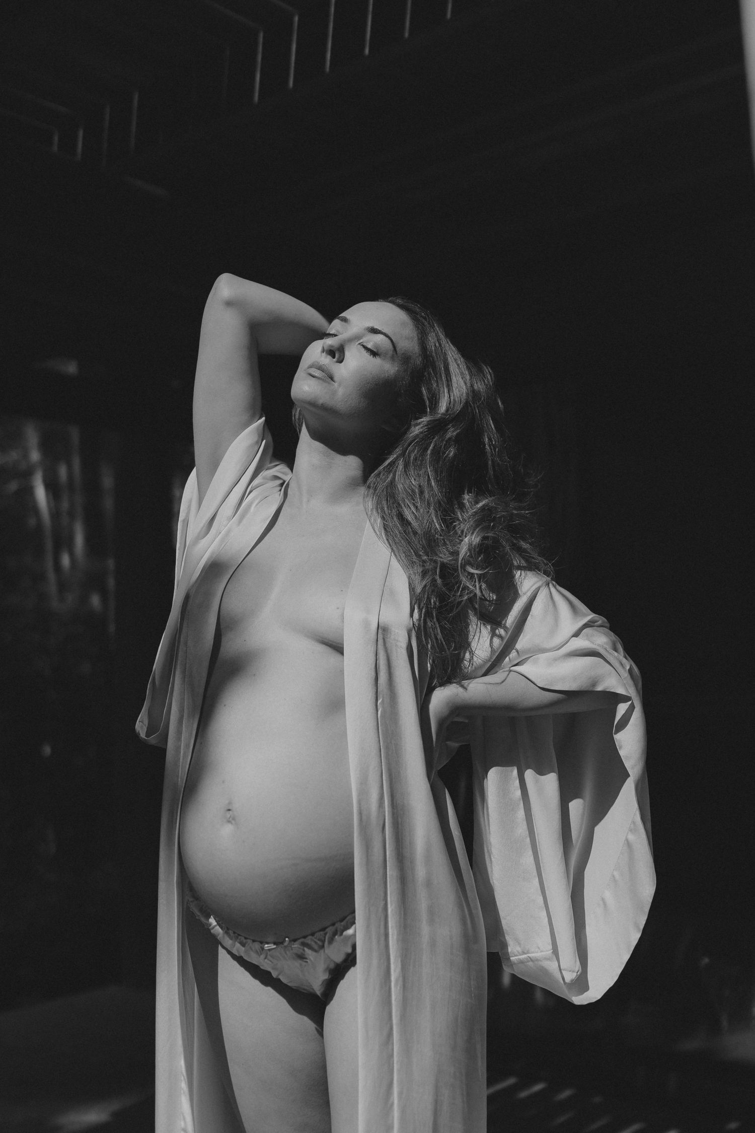 An artistic black and white maternity boudoir photo featuring soft natural lighting and flowing fabric.