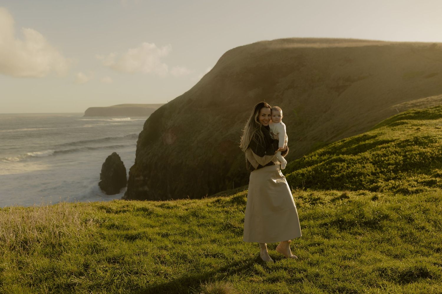 A woman holding a baby stands on a lush green coastal cliff with dramatic ocean views and golden sunlight.