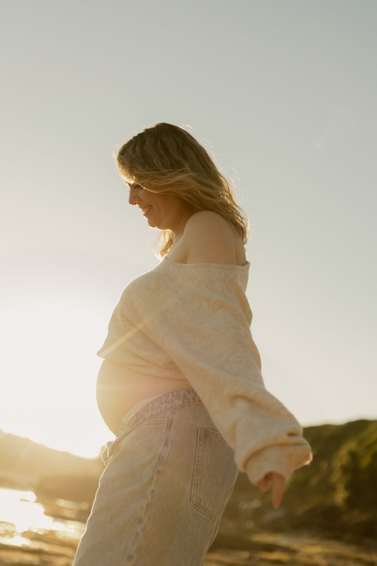Pregnant woman in cream sweater and jeans smiling at sunset on a beach, golden backlight glowing behind her.