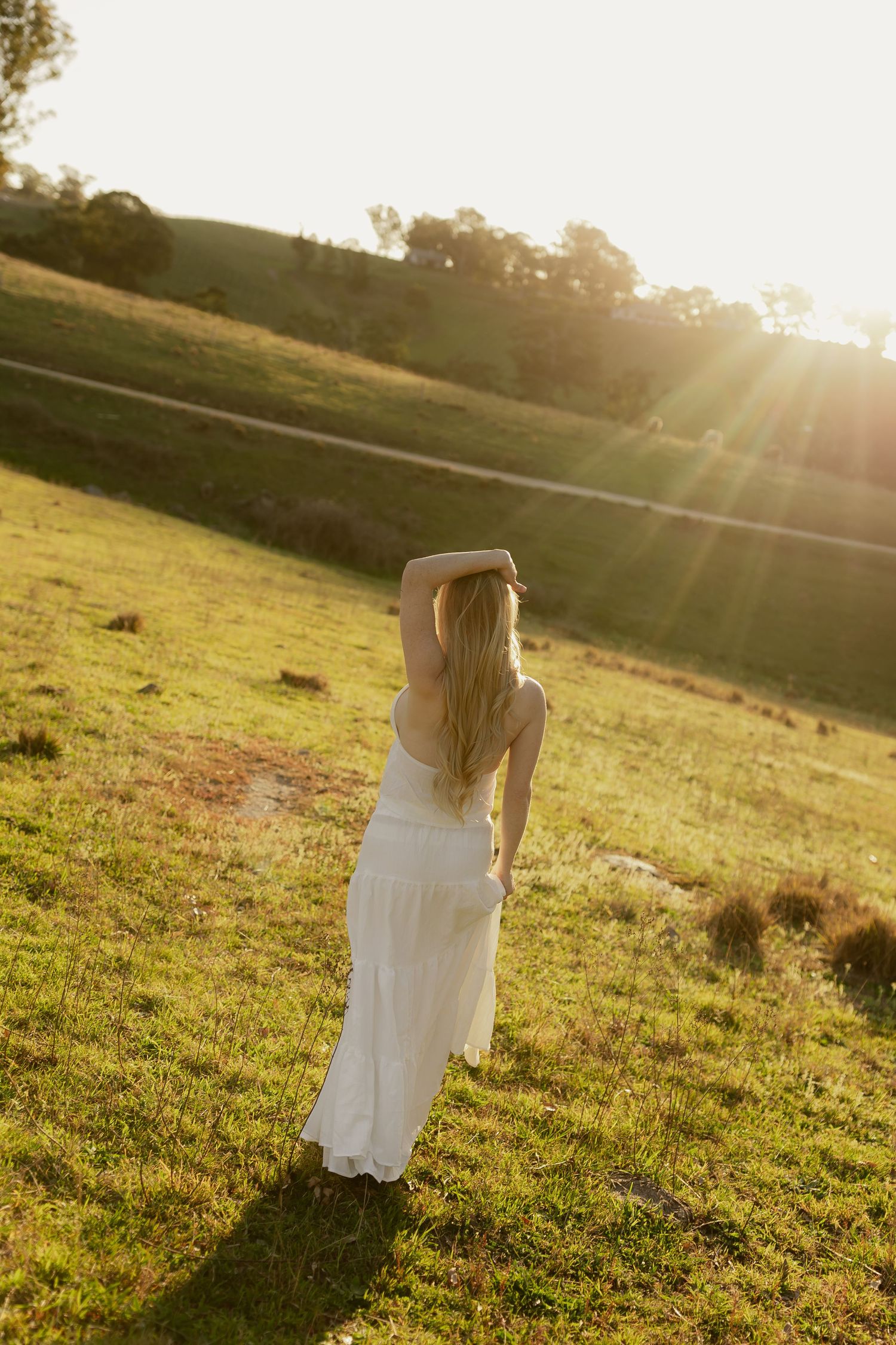 Blonde woman in white dress facing sunset with arms raised on lush green hillside.