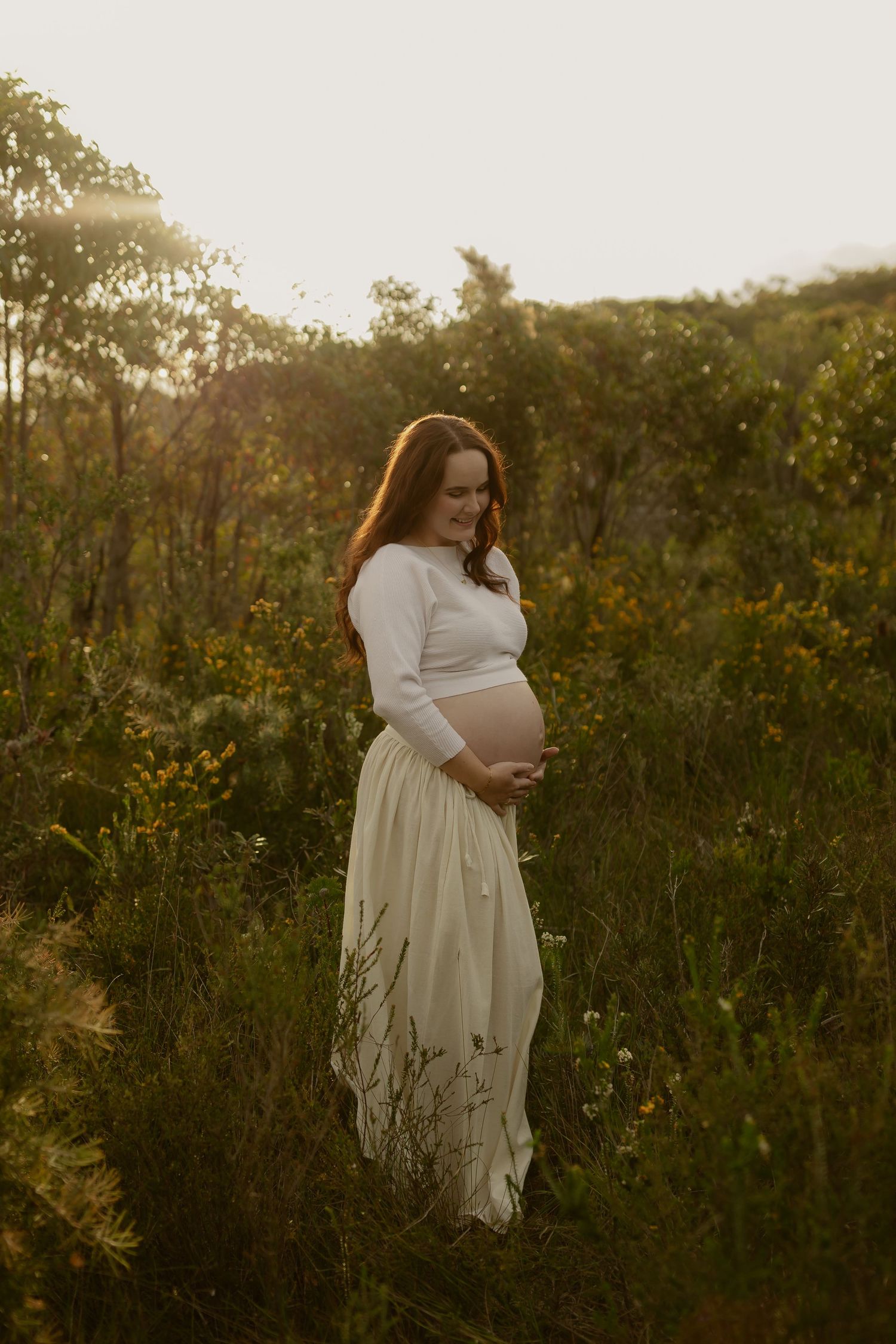 Pregnant woman in white crop top and skirt cradling baby bump in golden hour sunlit wildflower field.