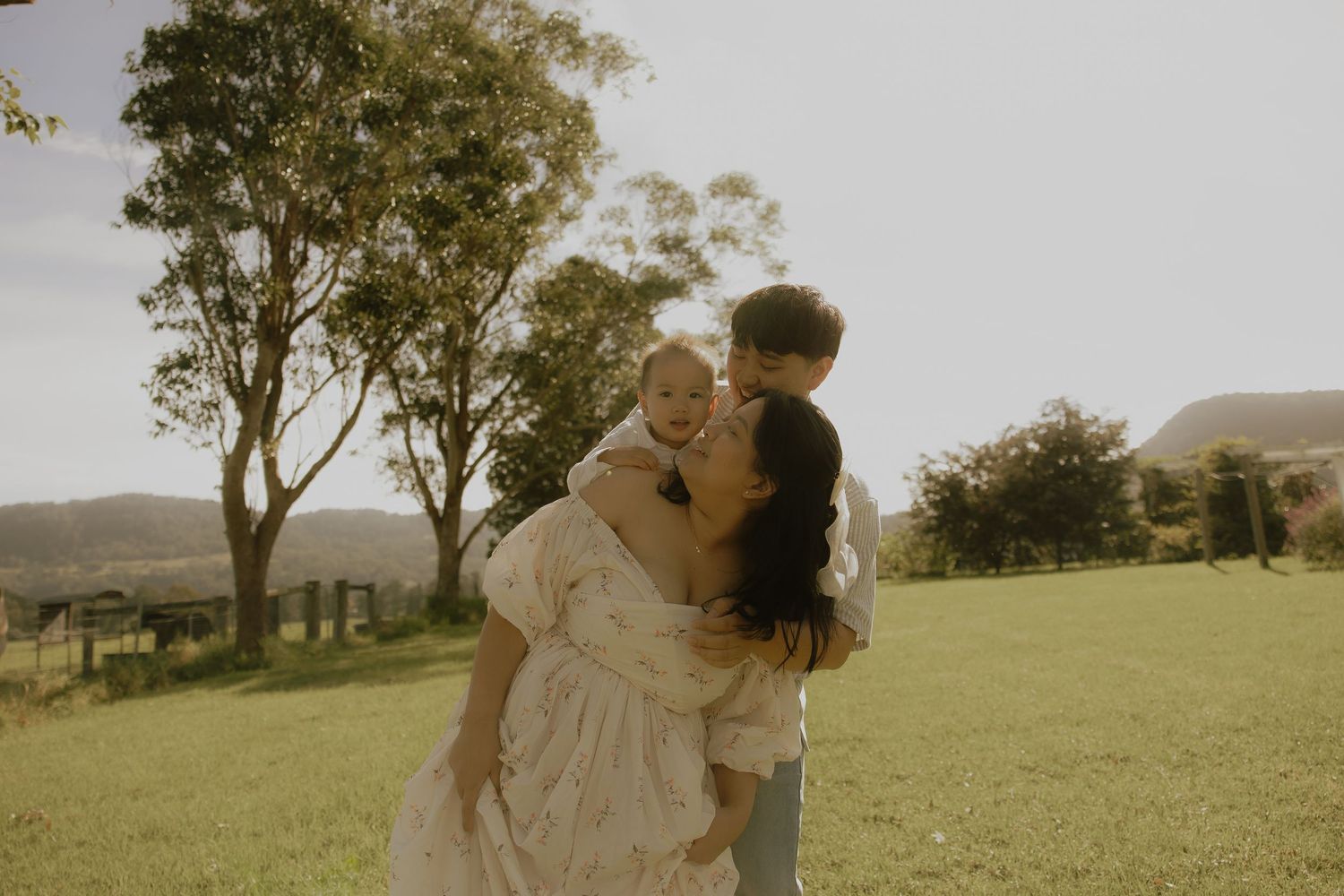 A mother holds her two young children in a sunlit green field with trees and mountains in the background.