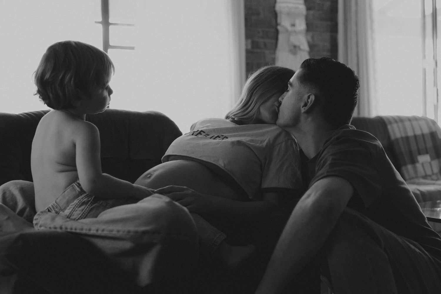 Black and white intimate photo of a pregnant couple kissing on a sofa while a toddler watches nearby.