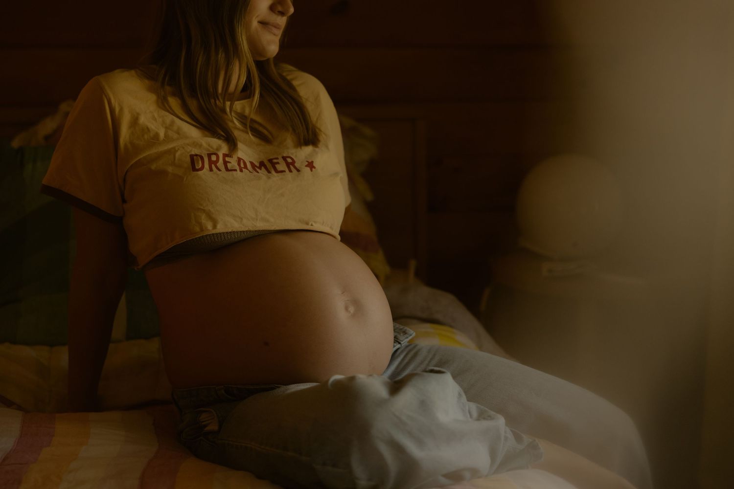 A pregnant woman in a yellow Dreamer crop top sits on a bed in warm soft light.