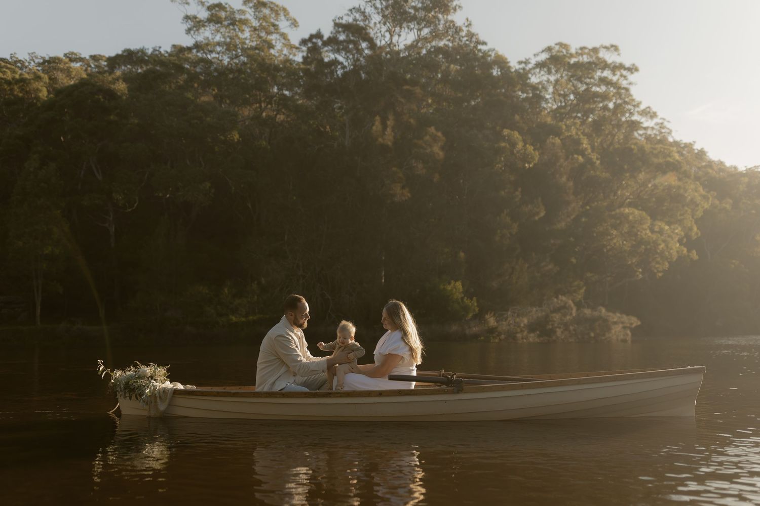 A family sits together in a white rowboat on a calm river surrounded by lush green trees in golden light.