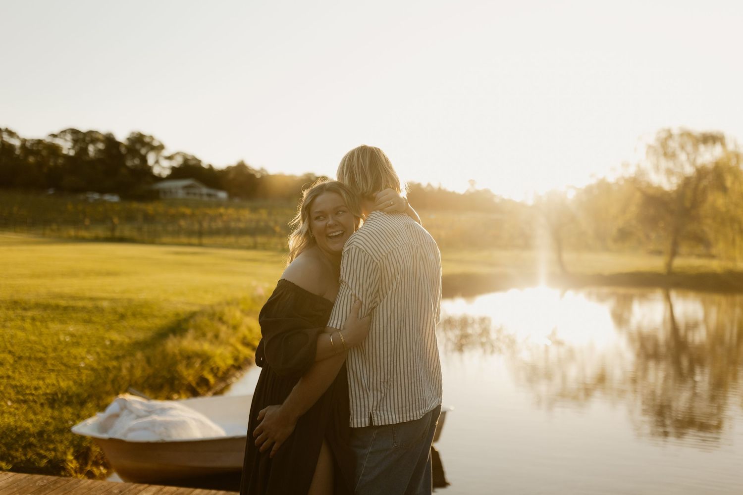 Couple embracing at golden hour by a serene pond, surrounded by lush greenery and warm sunlight.