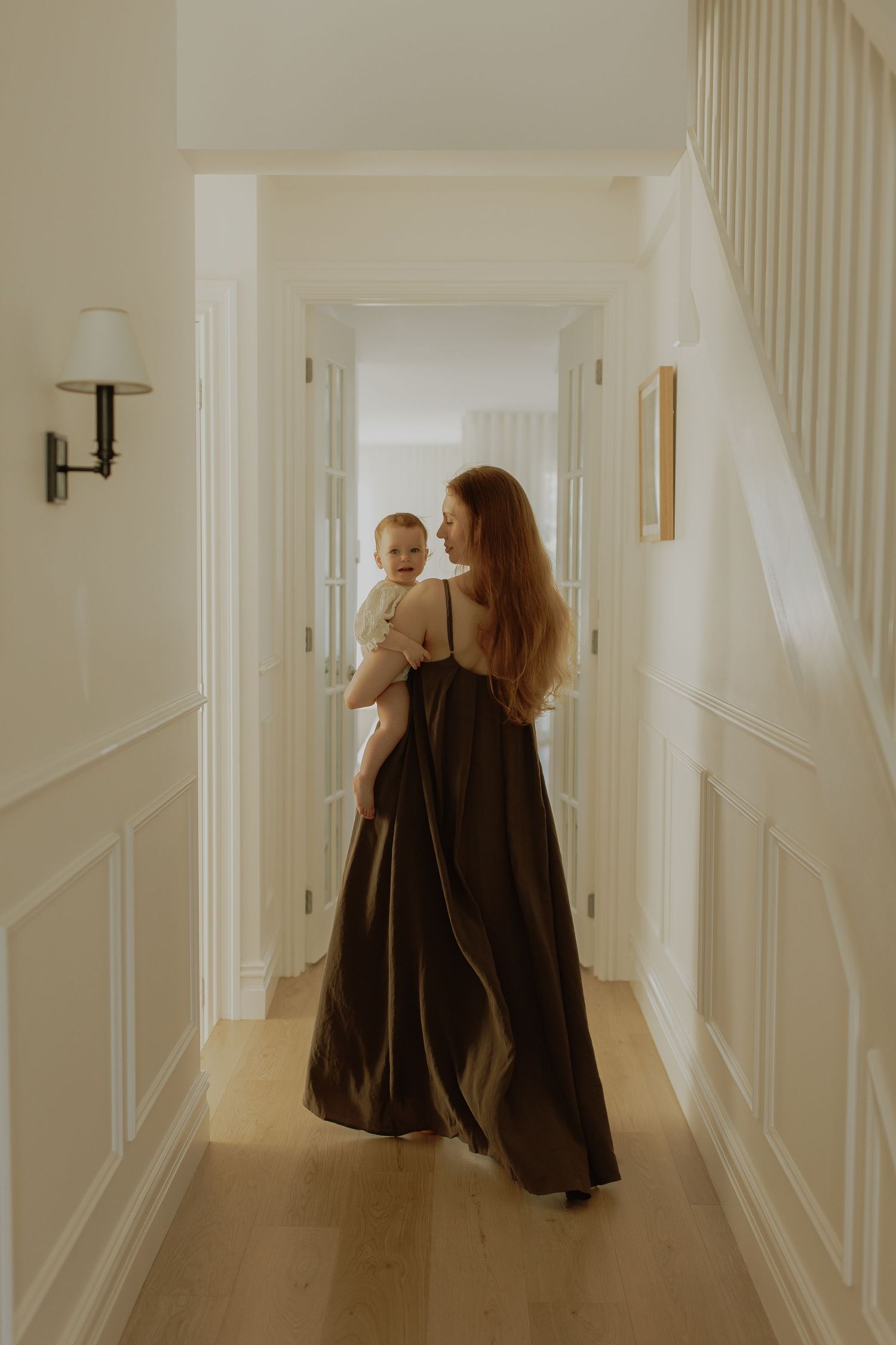 A woman in a dark maxi dress holds a baby while walking down an elegant white hallway with wood floors.