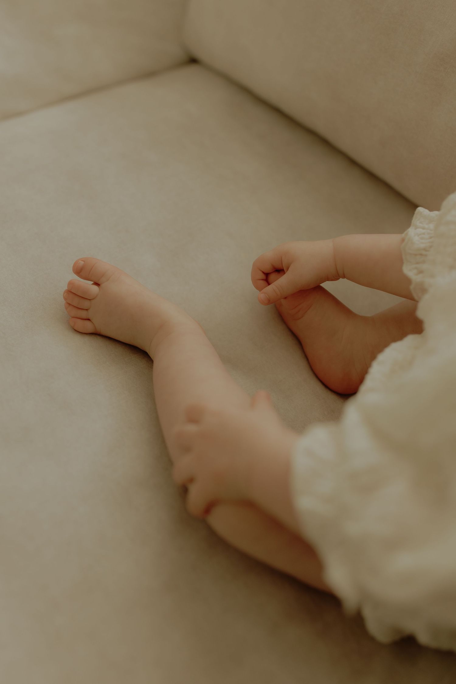Close-up of tiny baby feet and hands on a cream sofa, wearing a white lace outfit.