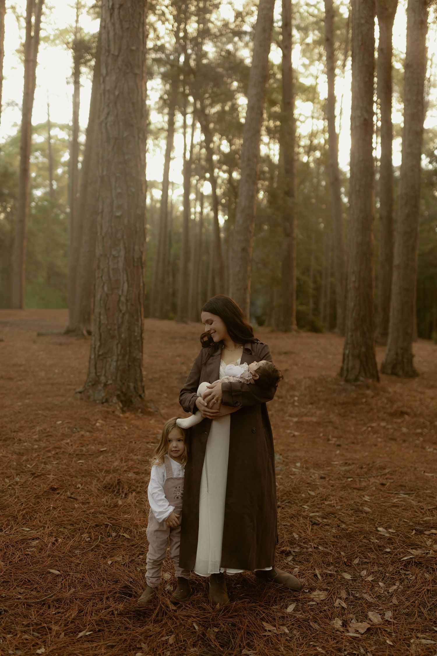 A woman in a white dress and dark coat holds a newborn while a toddler stands beside her in a pine forest.