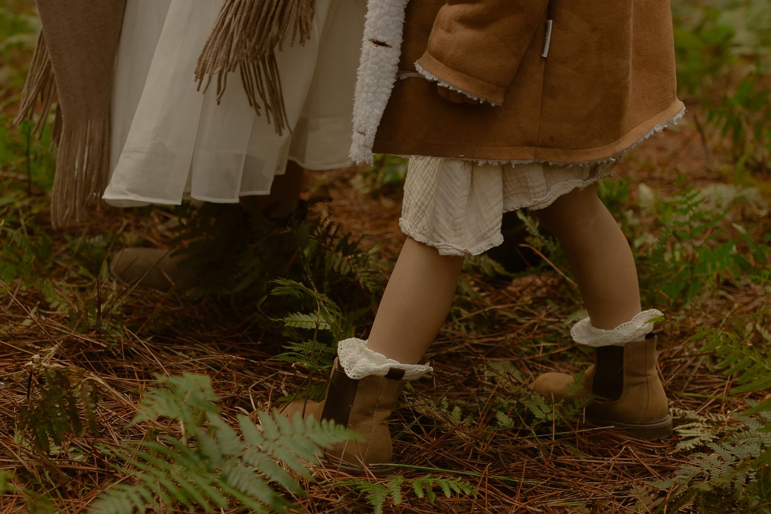 Two young children in boho-style dresses and brown ankle boots walking through a lush green forest setting.