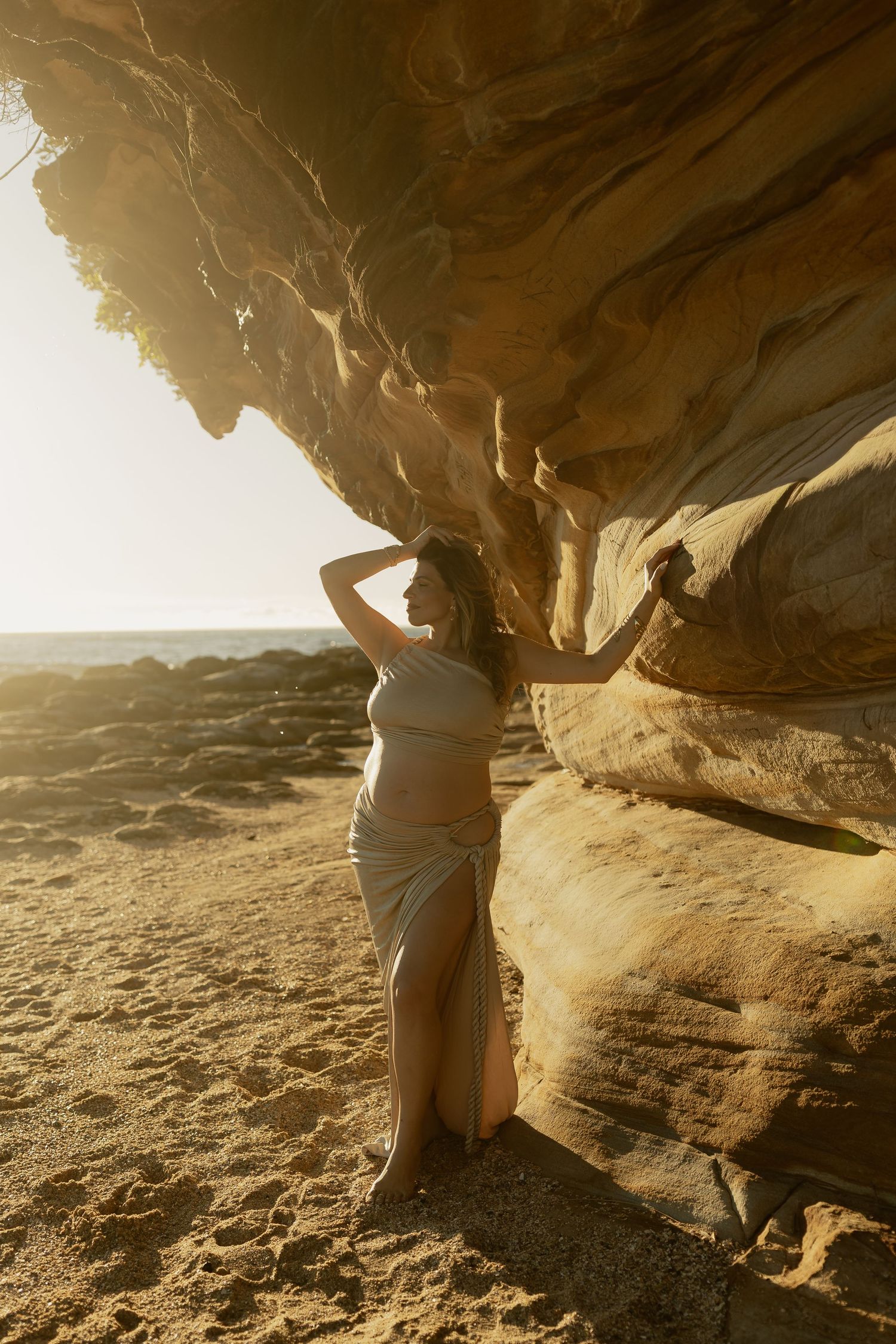 Pregnant woman in white dress poses against golden sandstone rock on sandy beach at sunset.