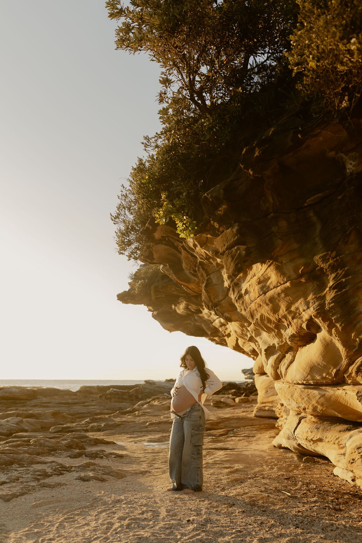 Pregnant woman in white top and gray pants standing by golden sandstone cliffs at sunset on a rocky beach.