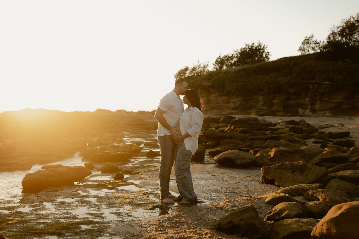 Couple sharing a romantic kiss on rocky beach at golden sunset, bathed in warm glowing light.