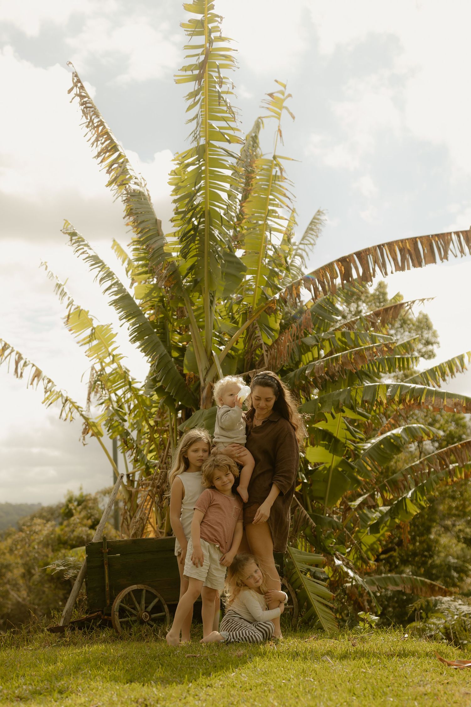 A mother holds her young children outdoors near a lush tropical banana tree on a bright cloudy day.