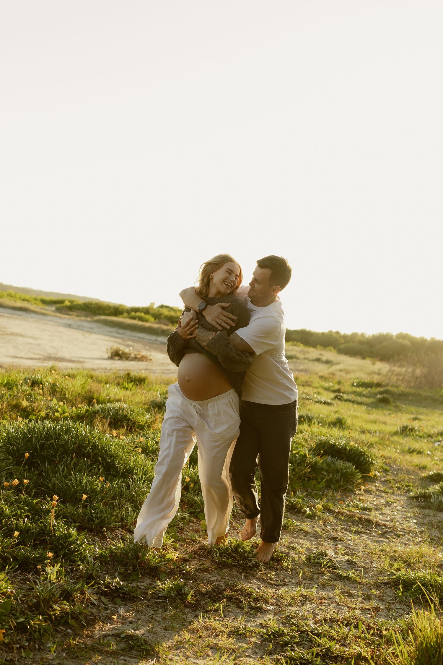 Happy couple embracing outdoors at sunset, pregnant woman laughing as partner hugs her from behind in green field.