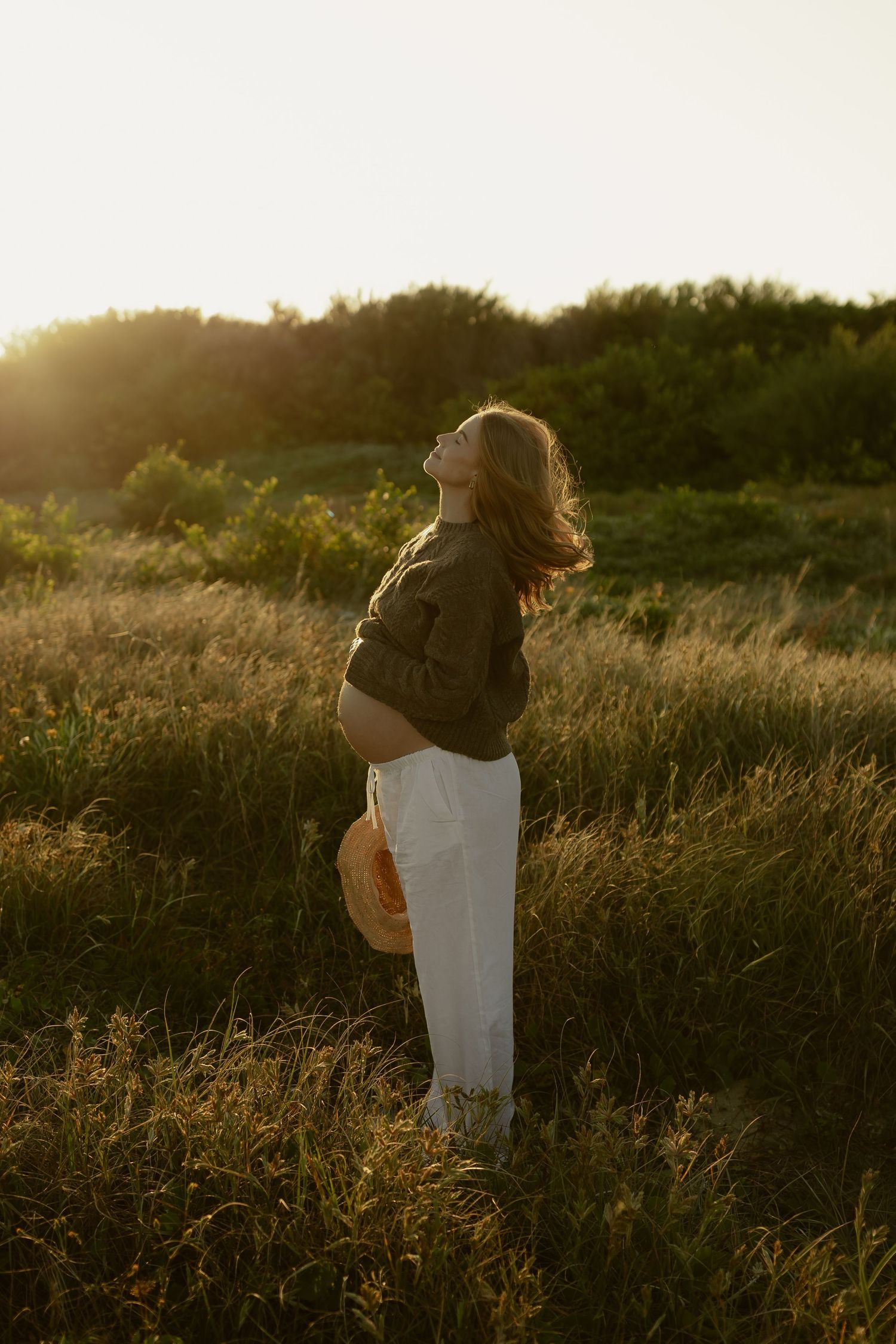 Pregnant woman in olive top and white pants holding straw hat while standing in golden sunset meadow.