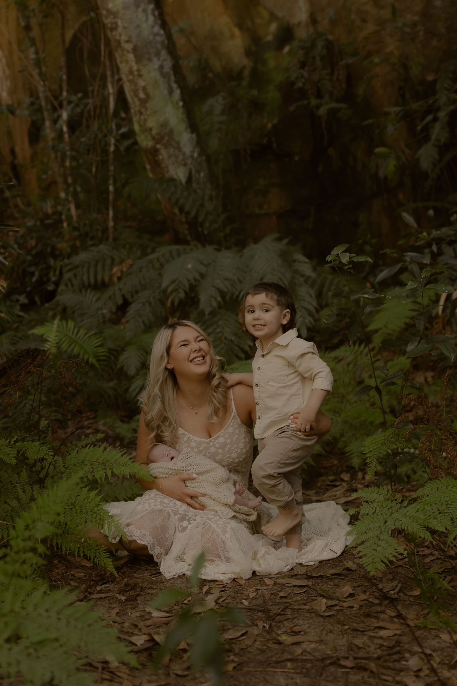 A laughing blonde woman in a lace dress sits with a smiling young boy in a lush fern forest setting.