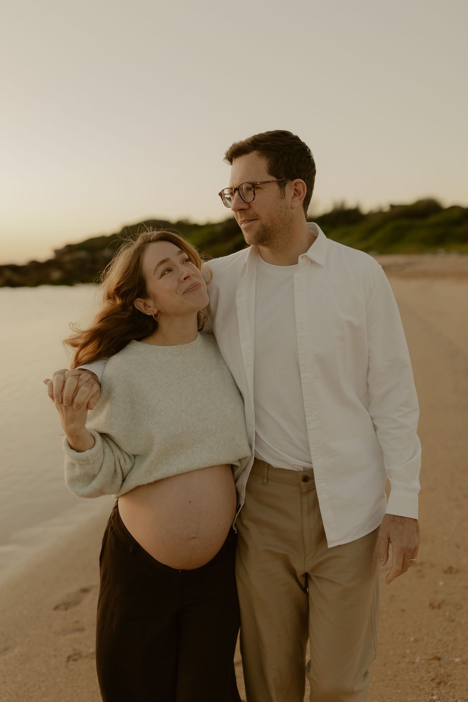 Pregnant couple embracing on beach at sunset, woman in gray sweater with baby bump, man in white shirt and glasses.