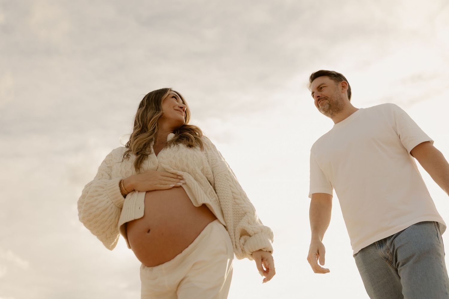 A smiling pregnant woman in a cream knit set holds her bump while walking with her partner on a beach.