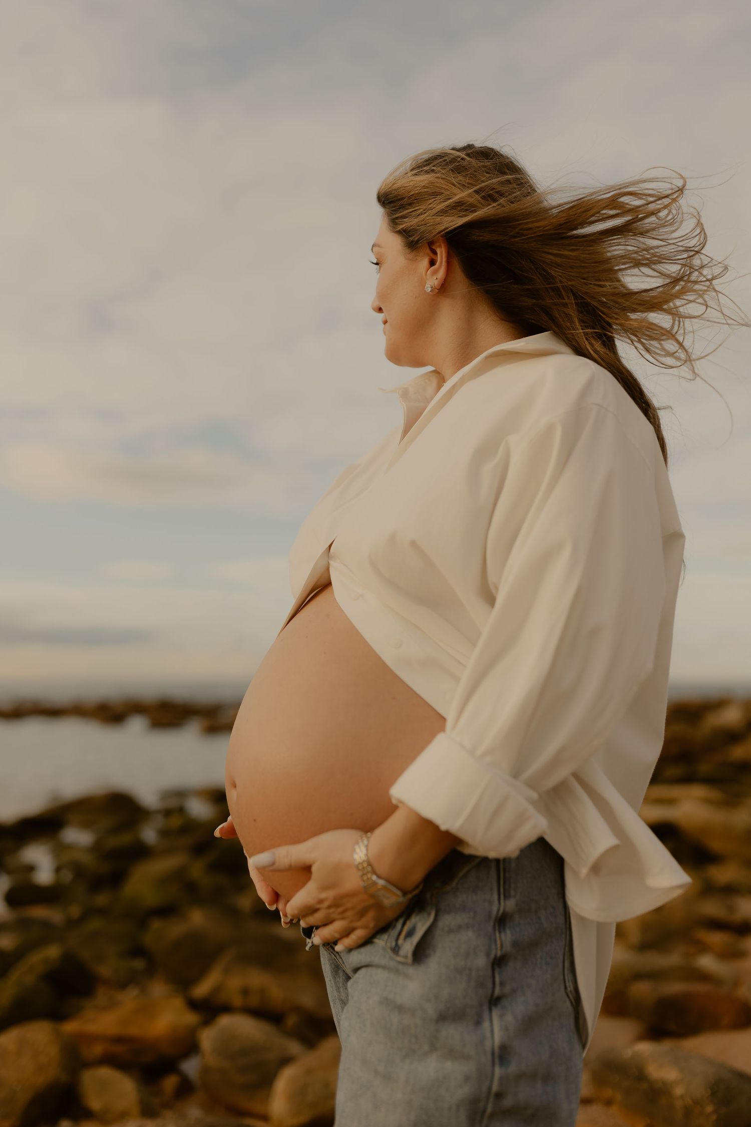 Pregnant woman in white shirt holding belly on rocky beach at sunset.