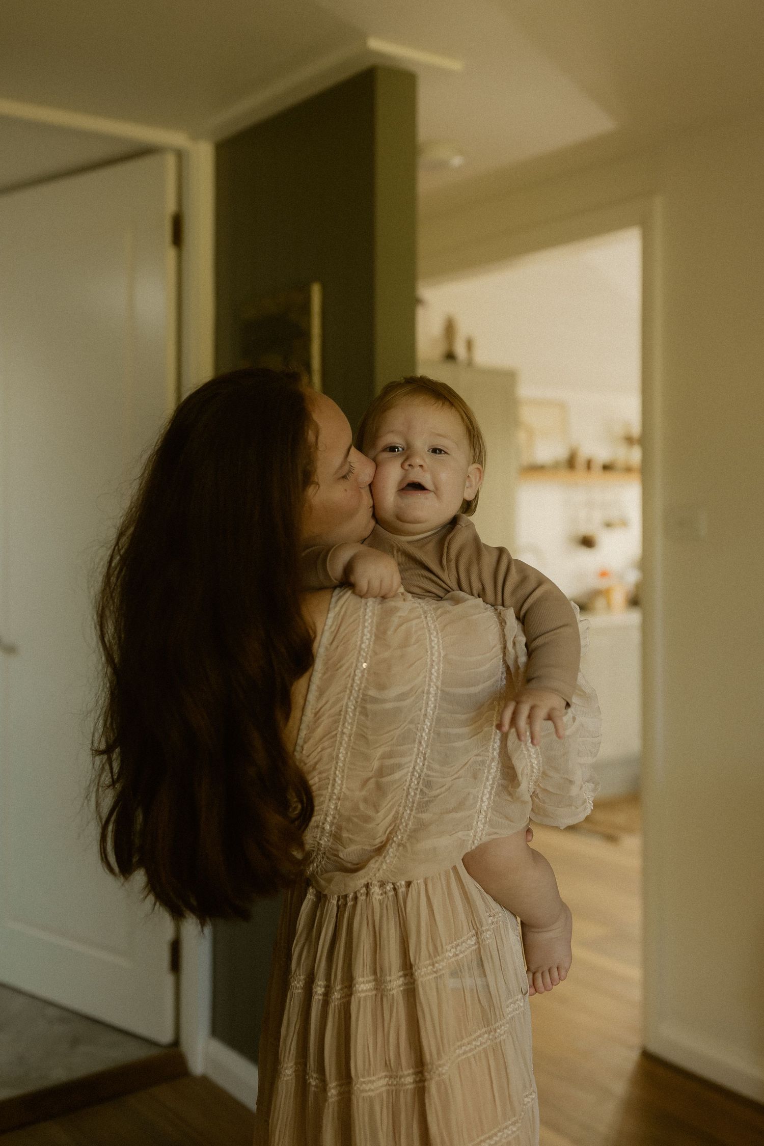 An intimate indoor portrait shows tender maternal affection in a light-filled home setting.
