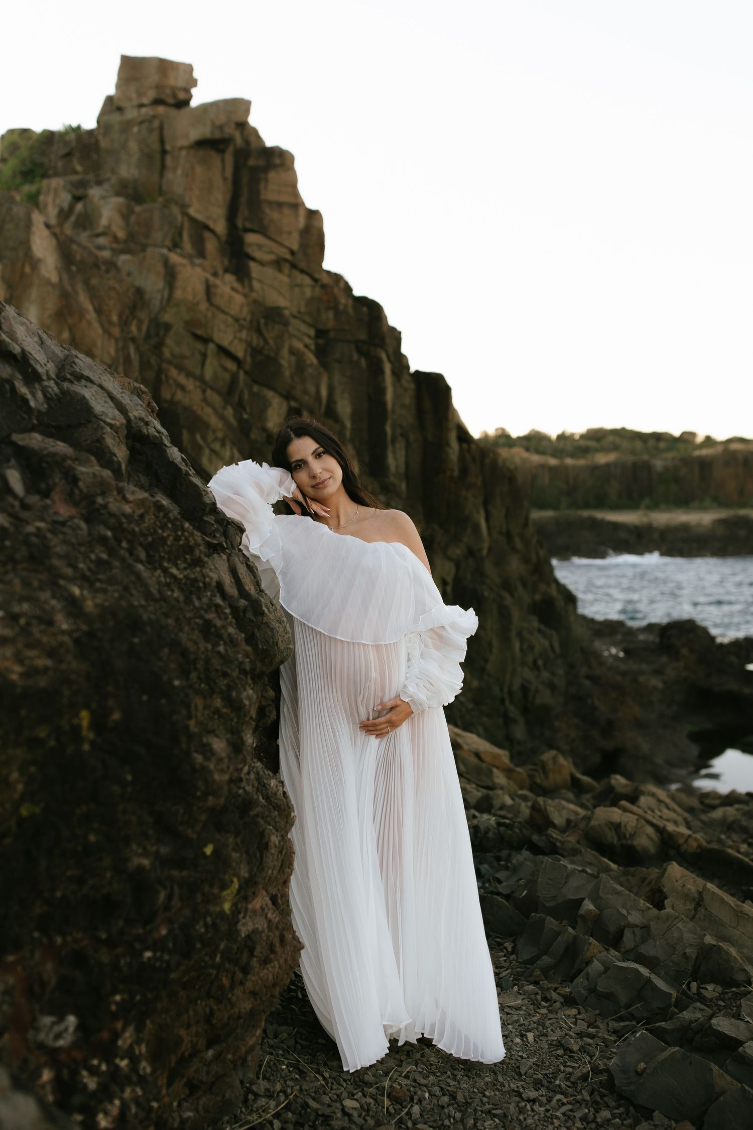 Ethereal white dress billows in the wind against dramatic coastal rock formations at sunset.