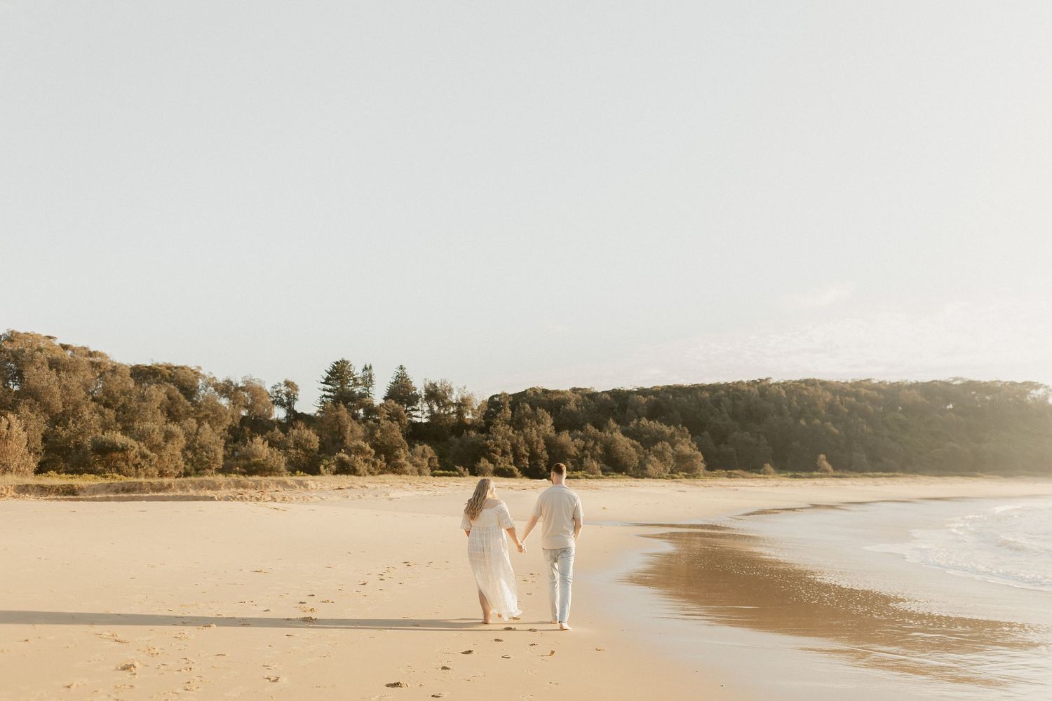 Couple holding hands walking on a sandy beach with lush green trees and calm ocean waves in golden light.