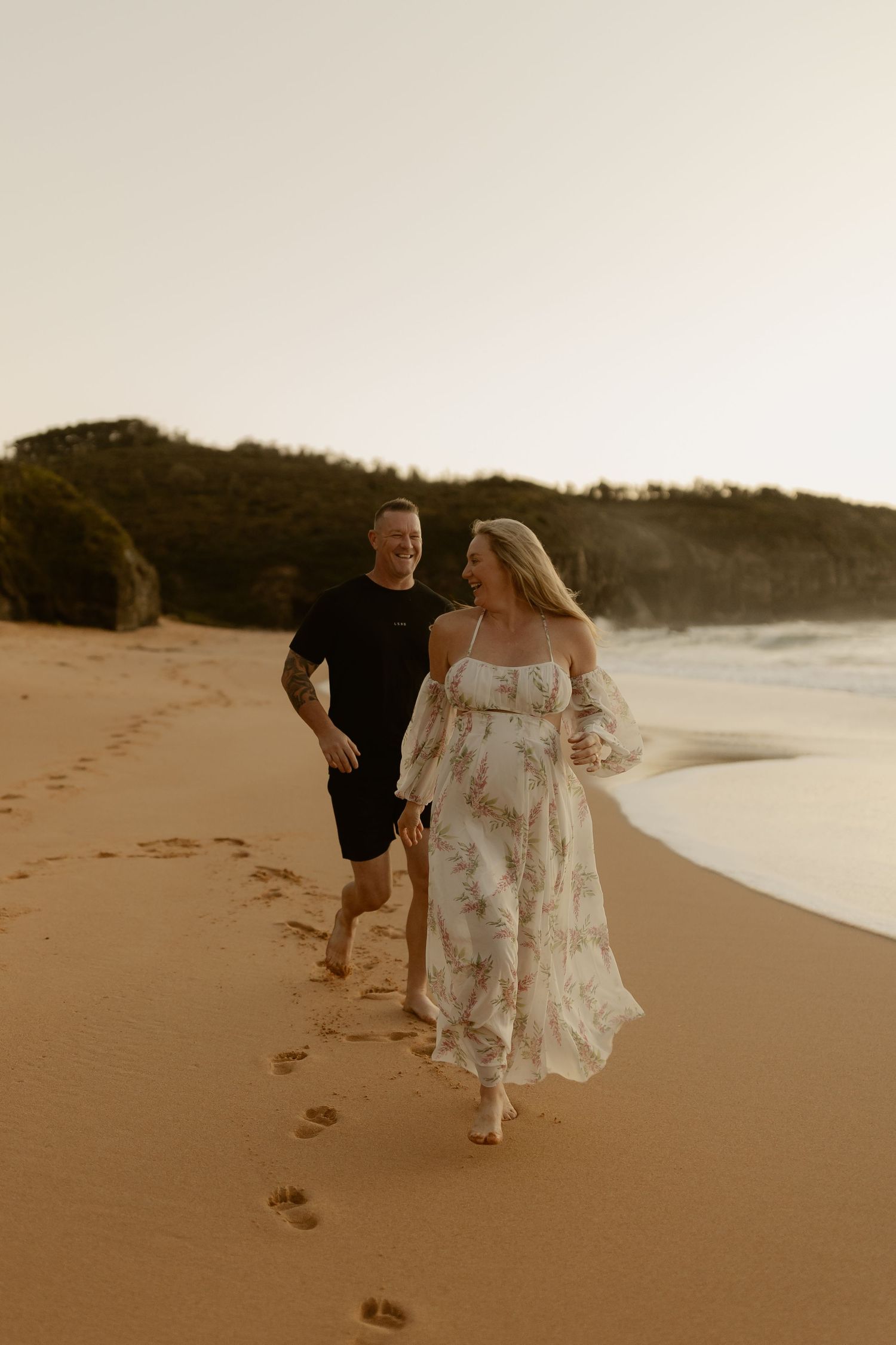 Pregnant couple walking barefoot on sandy beach at sunset, woman in floral off-shoulder dress, man in black outfit.