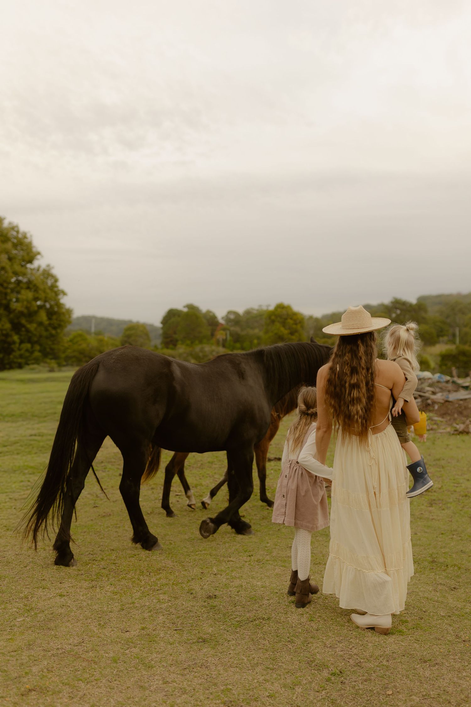 Woman in cream dress and sun hat walking with children alongside a black horse on a green farm.