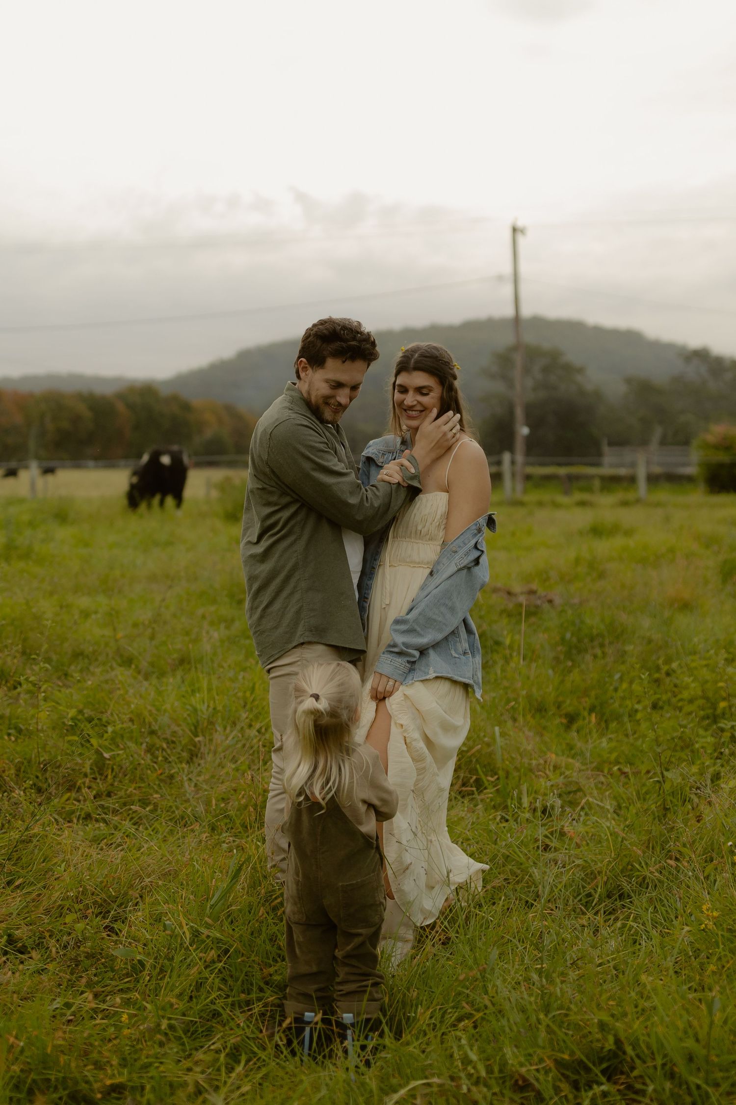 A couple laughs together in a green farm field while a young child stands between them on an overcast day.