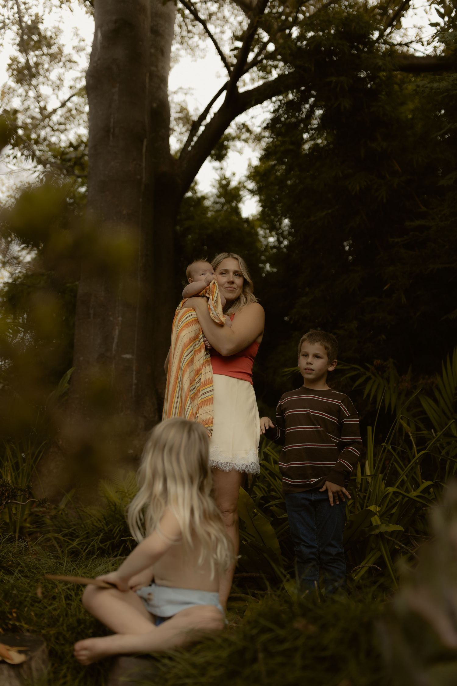 A mother holds a baby near trees while two children stand nearby in a lush garden setting.