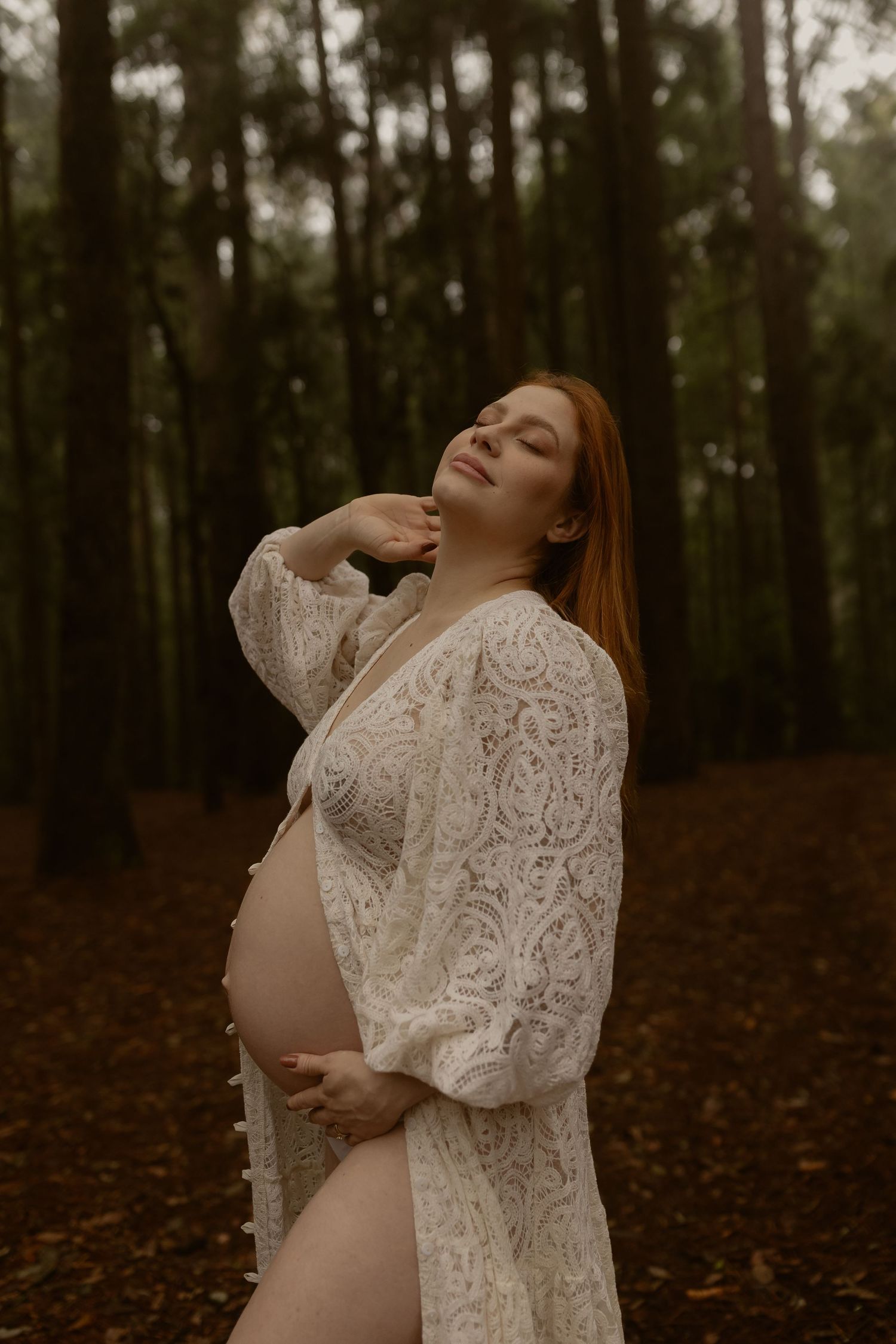Smiling pregnant redhead woman in white lace robe cradling her belly in an autumn forest.