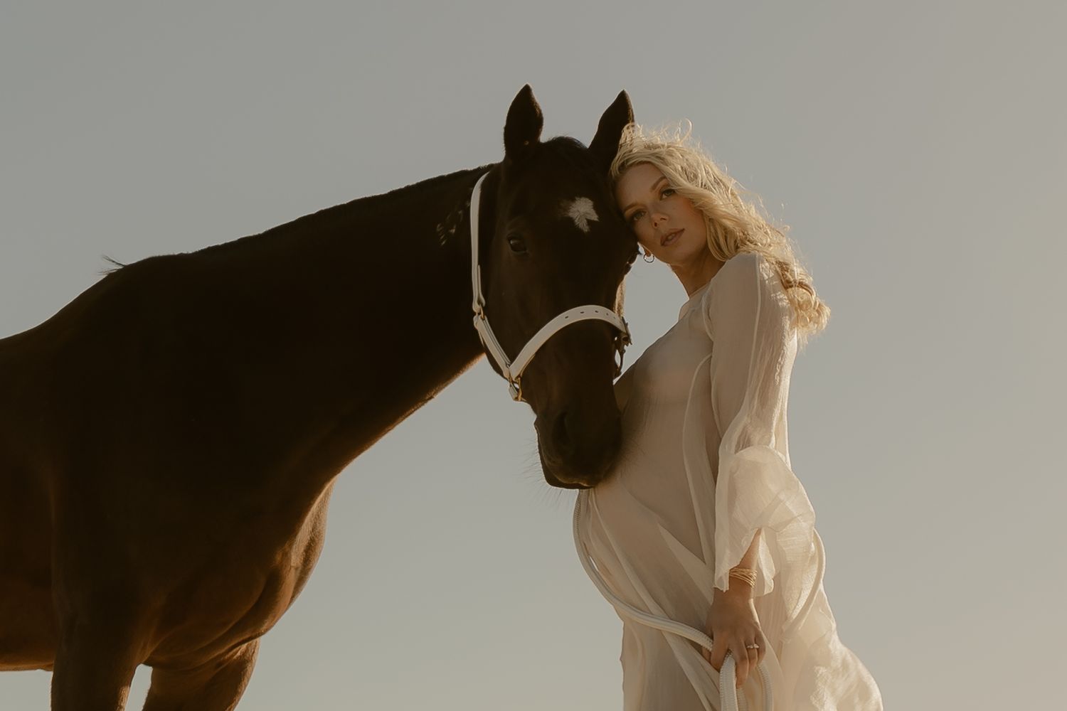 A blonde woman in a flowing white dress stands beside a dark brown horse against a soft sky backdrop.