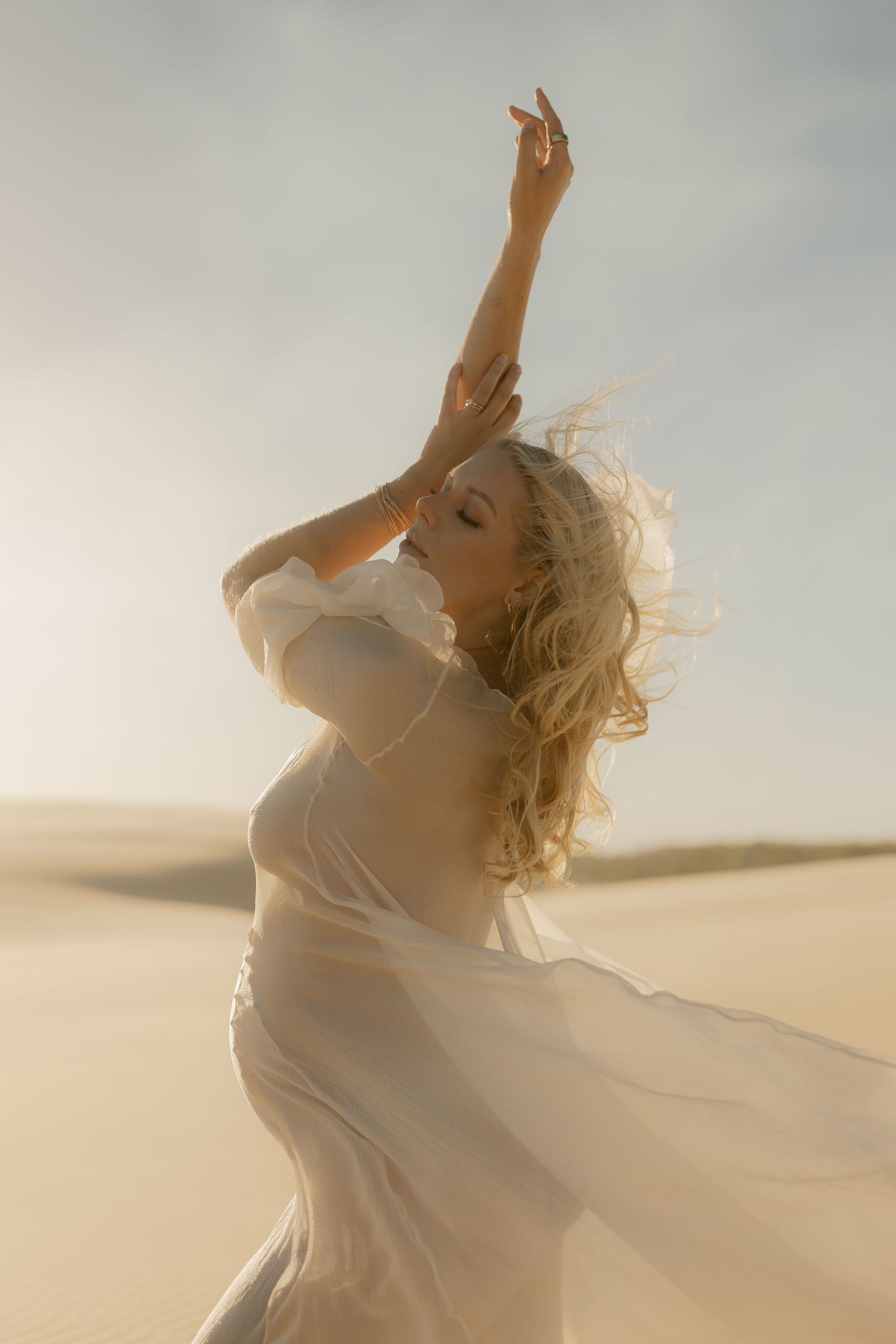 Blonde woman in sheer white dress with arms raised gracefully in a sunlit desert landscape.