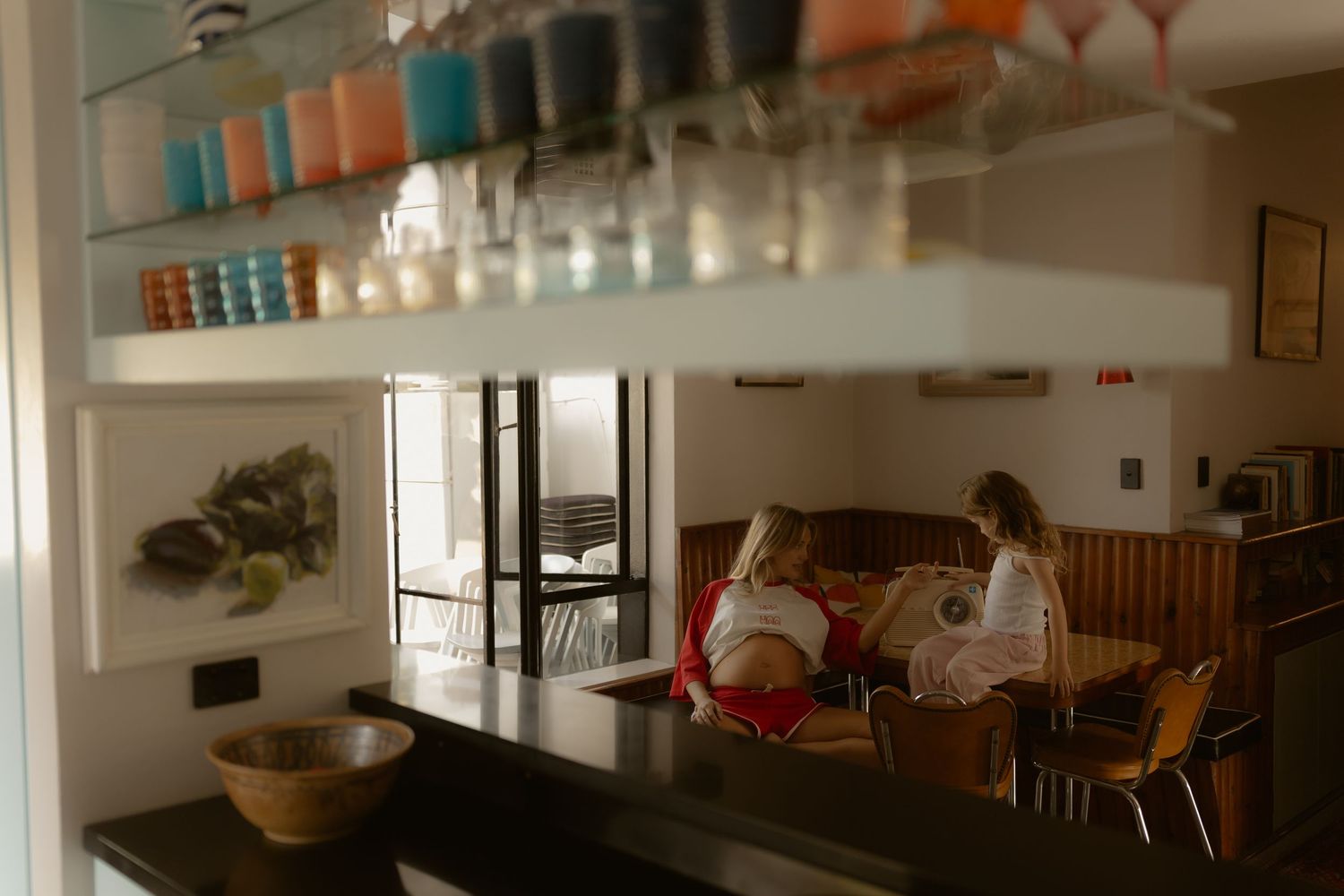 Two women sitting in a cozy restaurant booth beneath a shelf of colorful glassware and cups.