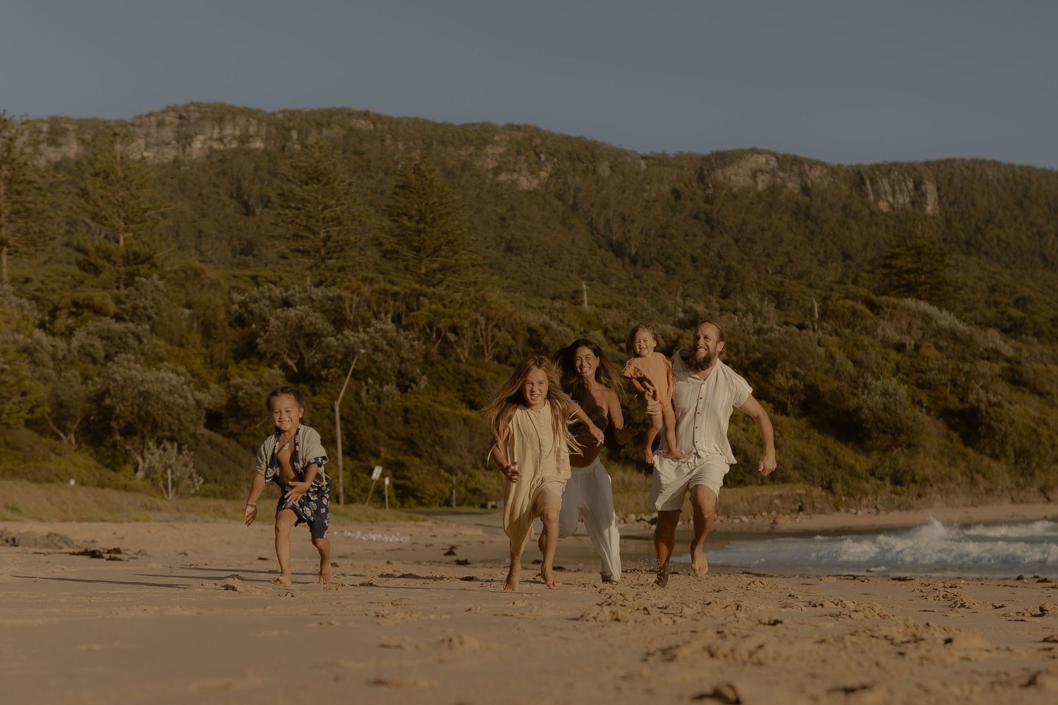 People walk along a scenic beach path with mountains and cliffs in the background during golden hour.