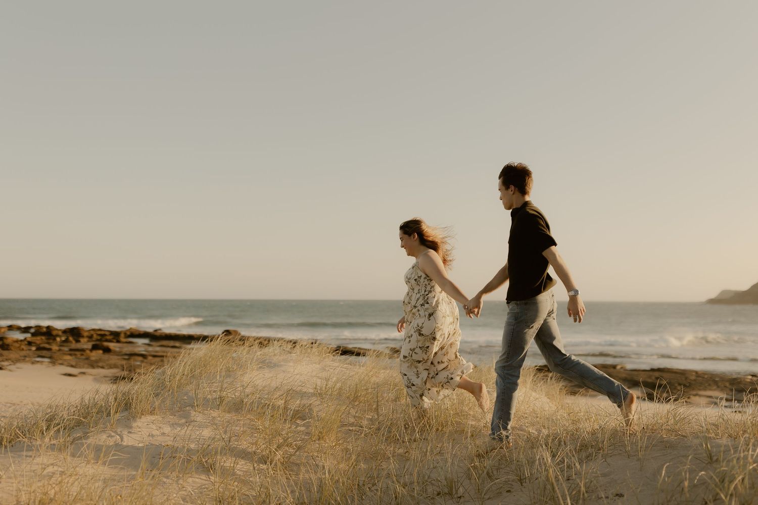 Two people run playfully through a golden field at sunset along the coastline, creating a dreamy silhouette.