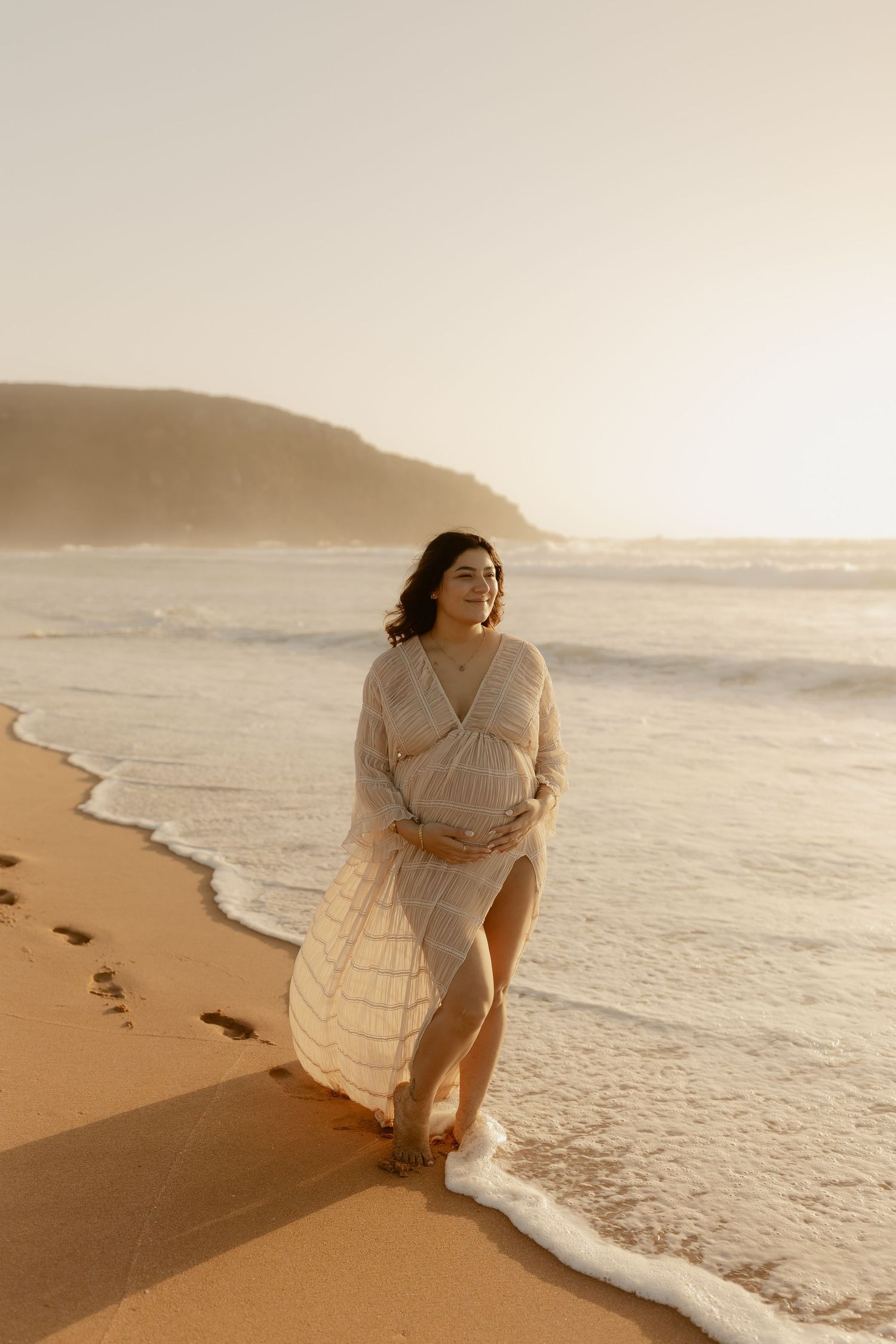 Person in white dress walks along sandy beach at golden hour leaving footprints in the sand.