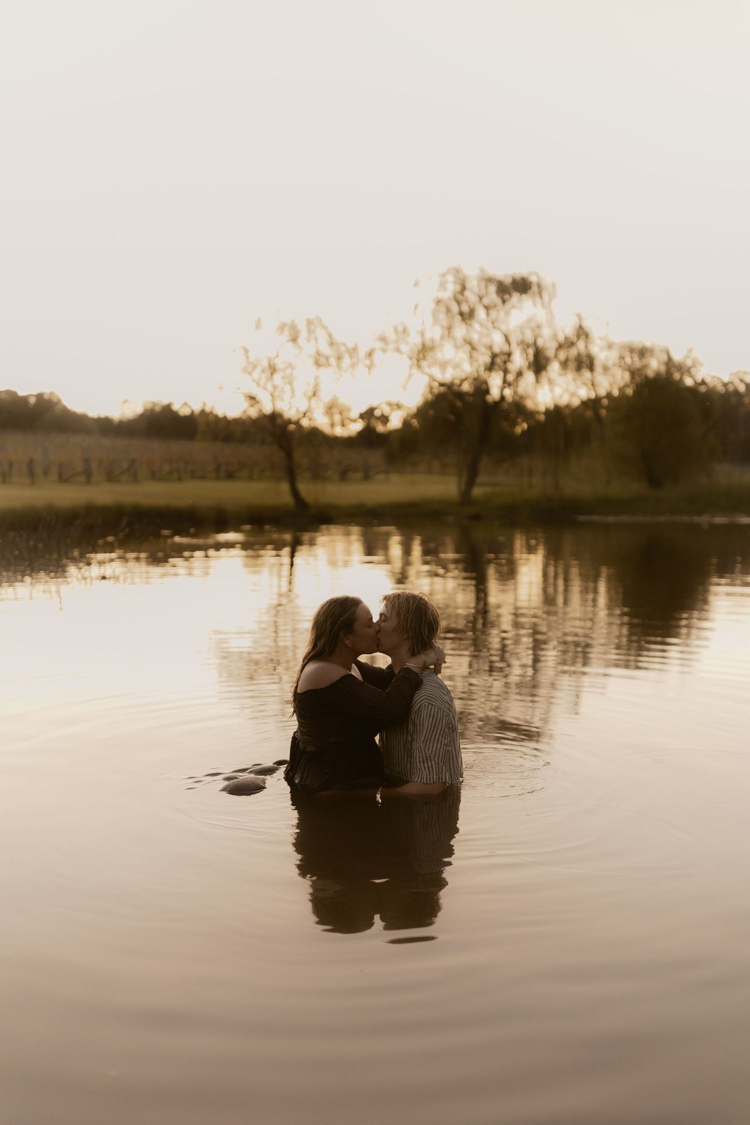 Two figures embrace while standing in still waters at golden hour with trees in background.