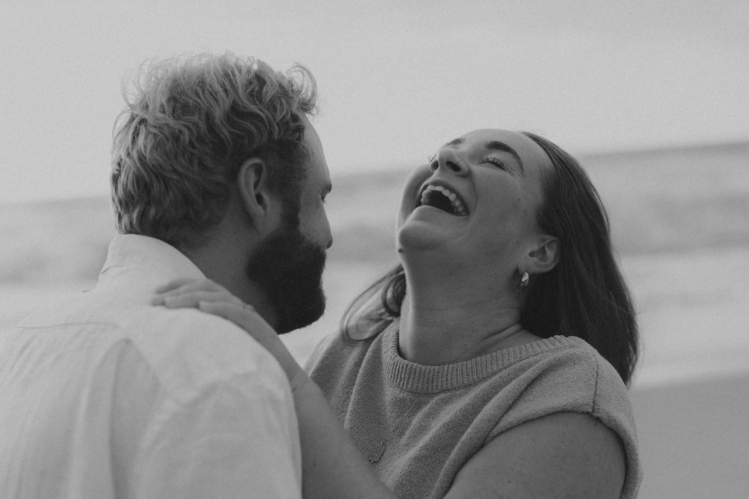 A romantic couple embraces and laughs together during a beach photoshoot.