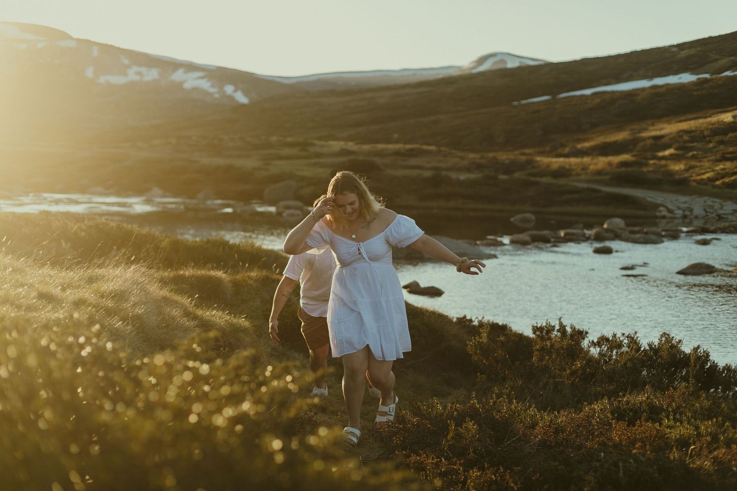 A person in a white dress walks along a coastal hillside path during golden hour sunset.