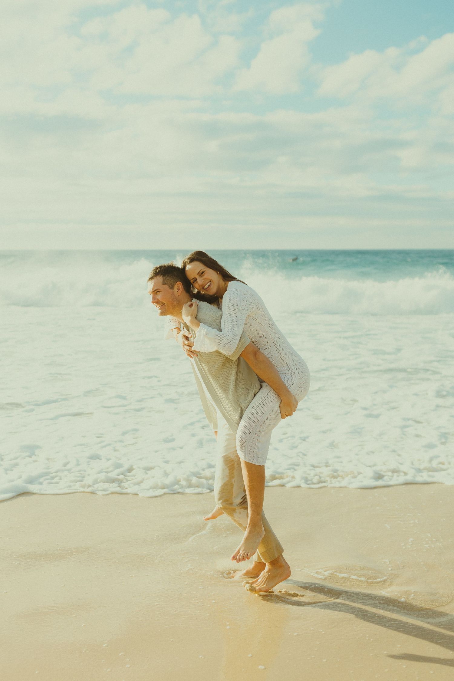 A playful piggyback ride moment between a couple in white and beige outfits on a sunny beach.