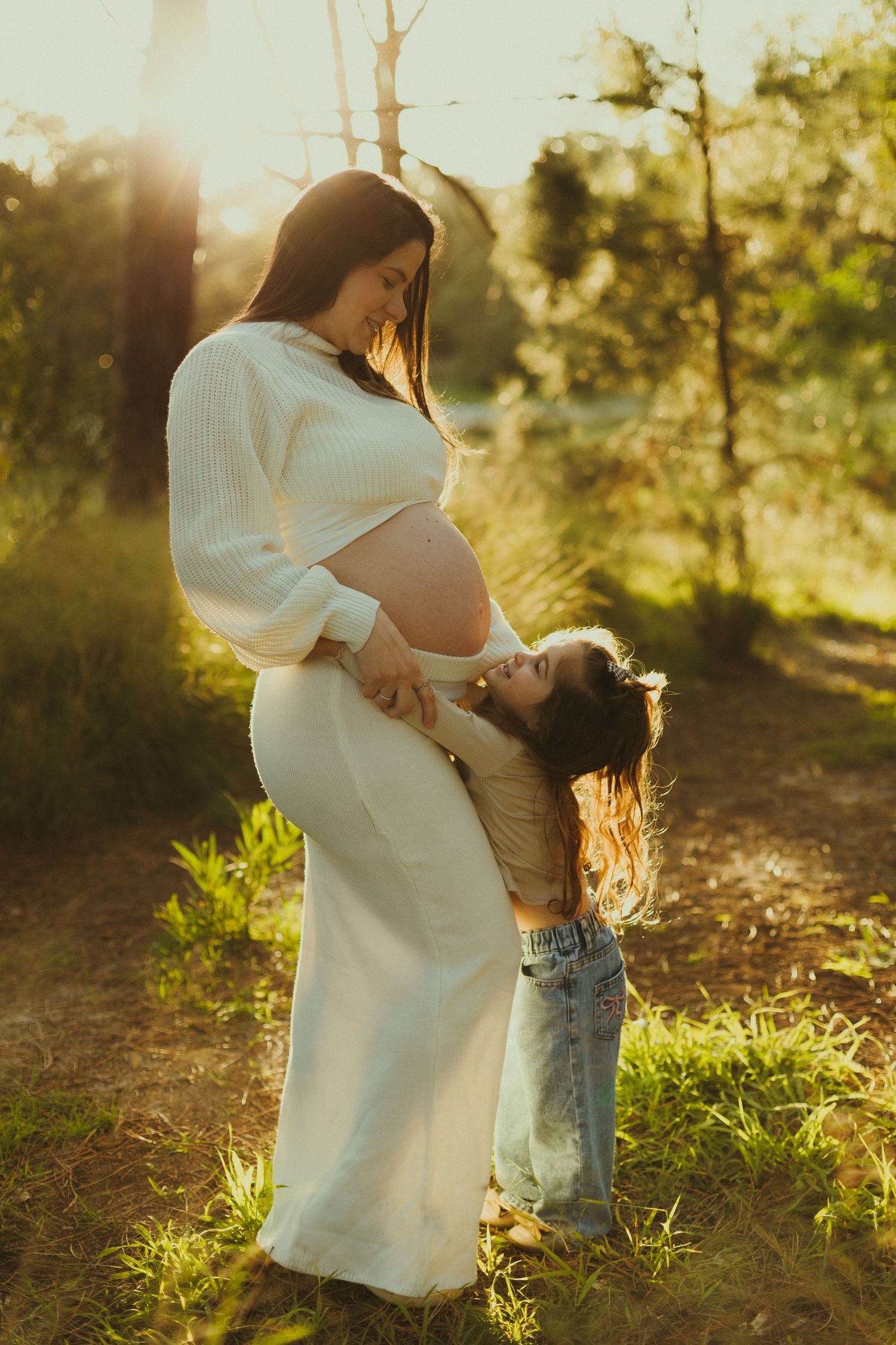 Child hugging expectant mother in white dress during golden hour photo session.