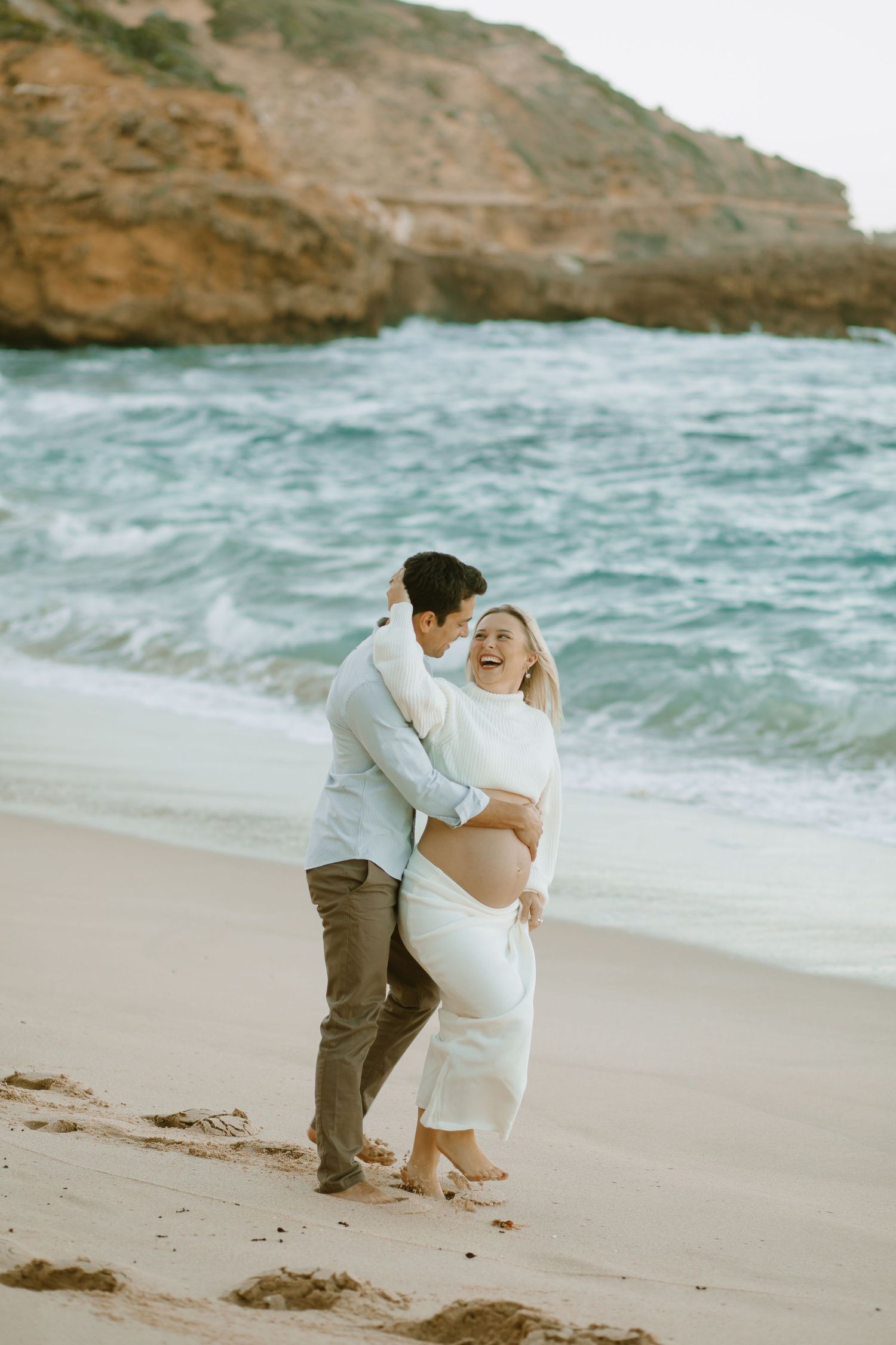 A couple embraces in a tender moment on a beach with dramatic coastal cliffs and turquoise waters.